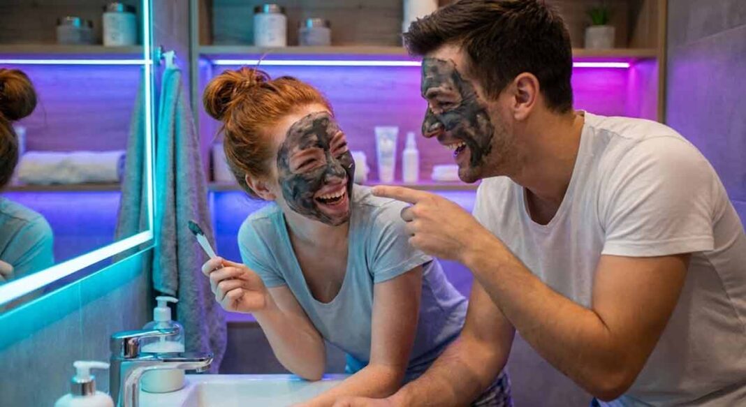 Polodetail na mladú ryšavovlasú ženu a muža v kúpeľni s fialovým a modrým LED podsvietením. Obaja majú na tvárach nanesenú tmavú pleťovú masku, smejú sa do nepríčetna a užívajú si spoločné domáce wellness rande.