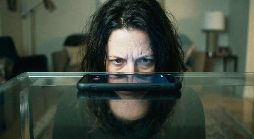 Detailný pohľad na ženu s podozrievavým výrazom, ktorej tvár osvetľuje žiariaci displej smartfónu položeného na sklenenom stole.