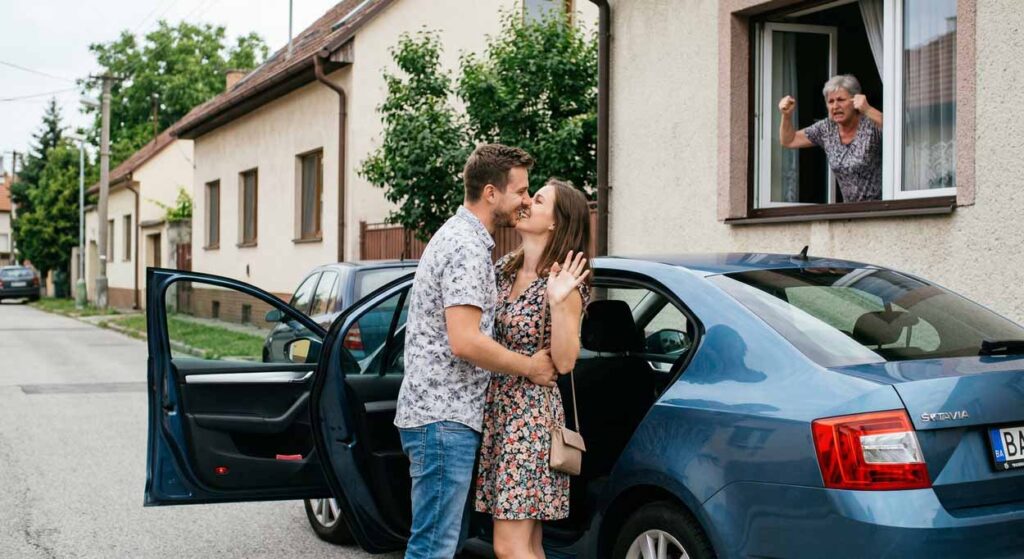 Mladý pár sa objíma pri aute, nahnevaná svokra kričí z okna domu, kontrolujúce správanie.