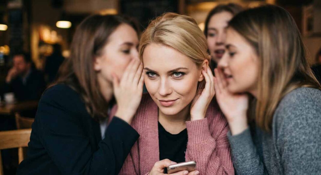 Detailný záber na tvár blonďavej ženy v ružovom svetri so zákerným úsmevom a šelmiími očami, ktorá so záujmom načúva šepkaniu drbov od svojich kamarátok v kaviarni. Ilustrácia psychológie manipulátorky a distribútorky ohovárania pre článok na portáli EXPOST.sk. Archetyp ohovárača.