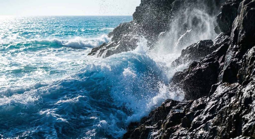 Detailný záber na kráľovsky modrú vlnu narážajúcu do tmavého skalnatého útesu, pri ktorom sa uvoľňuje jemná biela hmla morského aerosólu a milióny drobných kvapôčok vody.