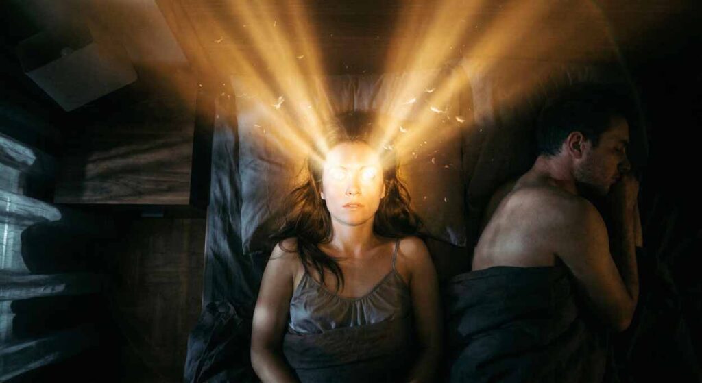 Žena ležiaca v tmavej spálni vedľa spiaceho muža, z jej očí vyžarujú dva silné lúče teplého svetla smerom k stropu, vizualizácia hyperaktívnej mysle a nespavosti v tichu.