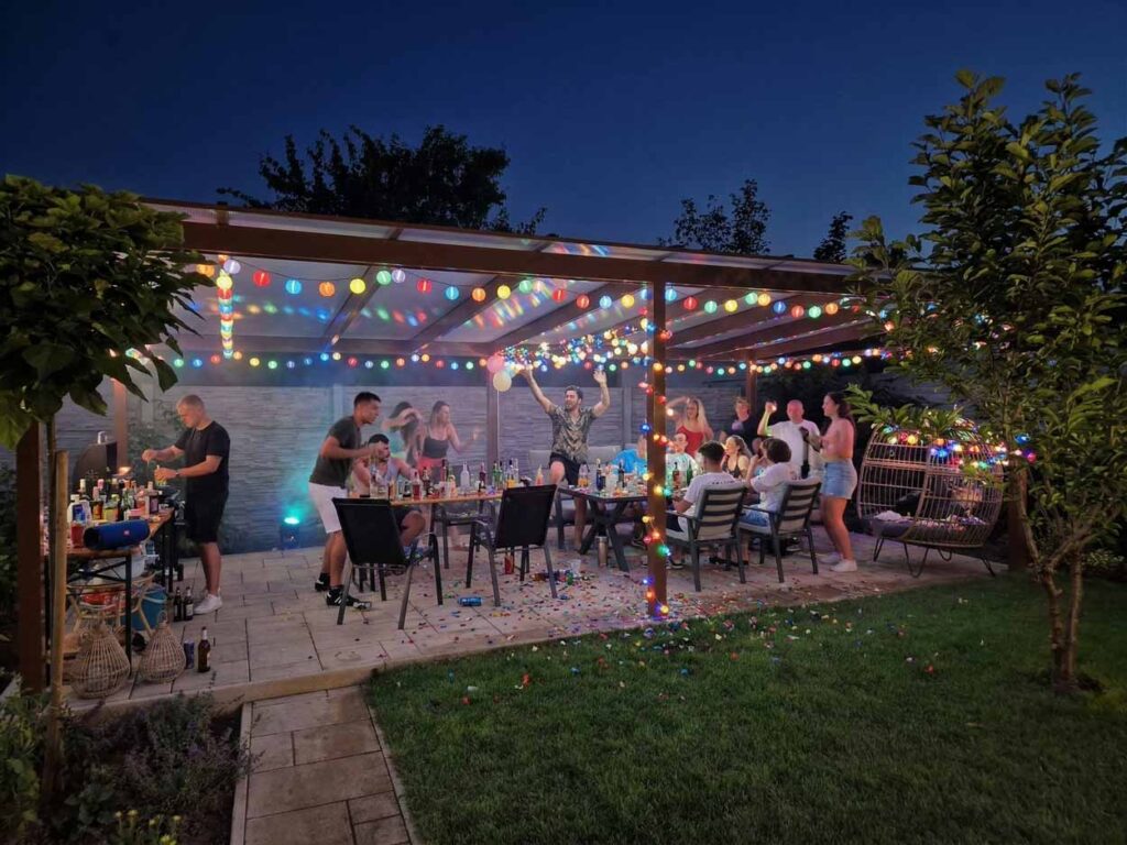 Nočná outdoorová párty na dvore pod prístreškom s farebnými svetelnými reťazami. Skupina ľudí je zhromaždená okolo stola. Muž na ľavej strane pripravuje nápoje pri barovom pulte.