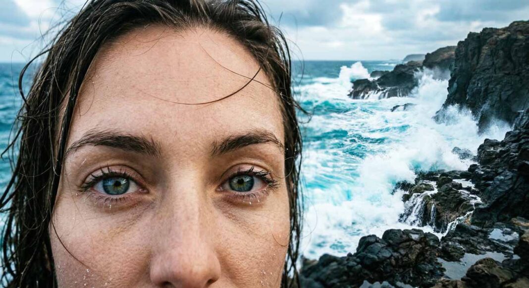 Detail pokojných očí človeka hľadiaceho na azúrové vlny mora trieskajúce o útesy, zatiaľ čo cez dramatické mraky prenikajú rovnobežné lúče slnka.