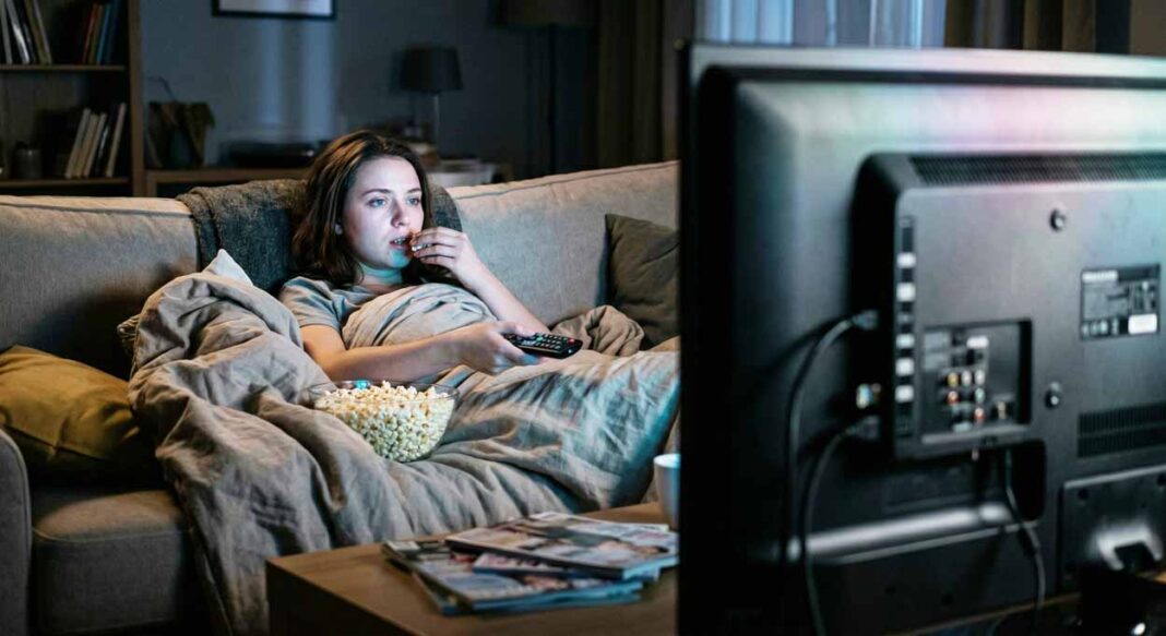 Žena v tmavej obývačke pod perinou na gauči unavene tupo pozerá na TV, s pukancami.