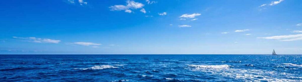 Rozsiahly výhľad na sýto kráľovsky modrý oceán, ktorý sa stretáva s jasnou kráľovsky modrou oblohou s roztrúsenými bielymi mrakmi počas slnečného dňa. V diaľke na obzore je vidieť malá plachetnica.